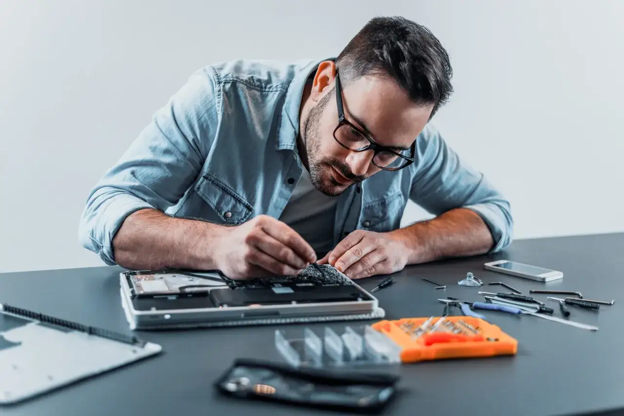 Mężczyzna w okularach naprawia laptopa, zastanawiając się, jakie parametry powinien mieć dobry laptop. Na stole narzędzia i części.