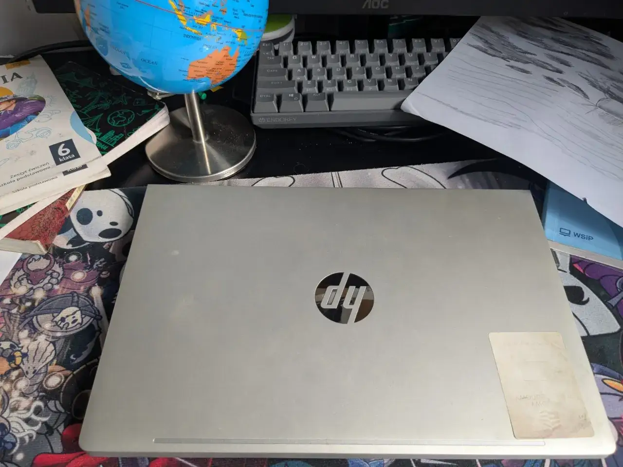 Na biurku leży zamknięty laptop HP, obok niego podręcznik do 6 klasy, globus i rysunek. Czy uczniowie dostaną laptopy?