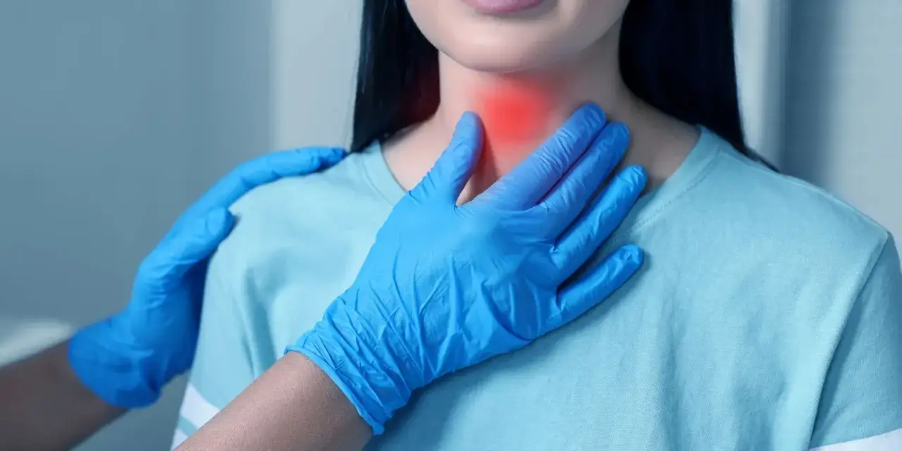 Lekarz w niebieskich rękawiczkach bada szyję pacjentki, gdzie widoczny jest czerwony obszar b&oacute;lu. T3 i T4 to hormony tarczycy.