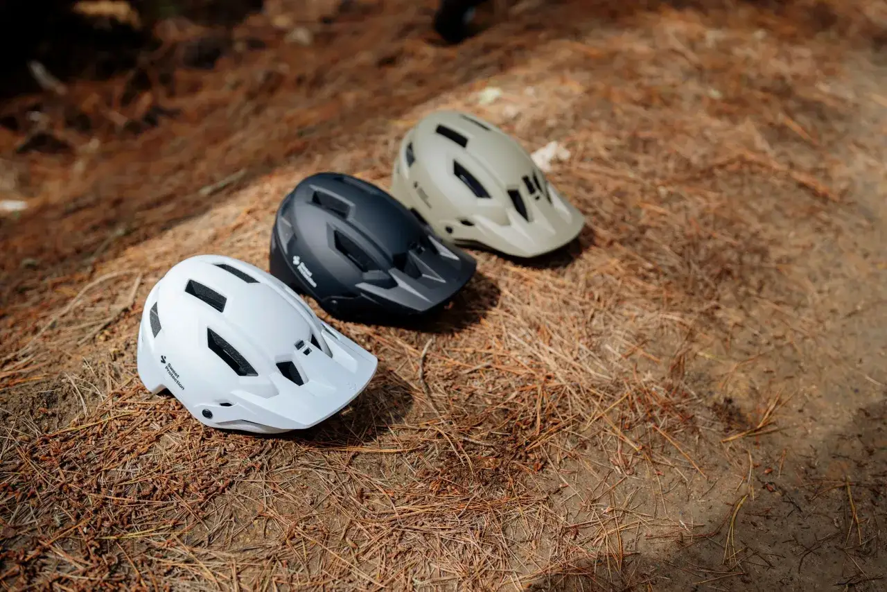 Trzy kaski rowerowe: biały, czarny i beżowy, leżą na leśnym poszyciu. Idealny wyb&oacute;r, jaki kask w g&oacute;ry?