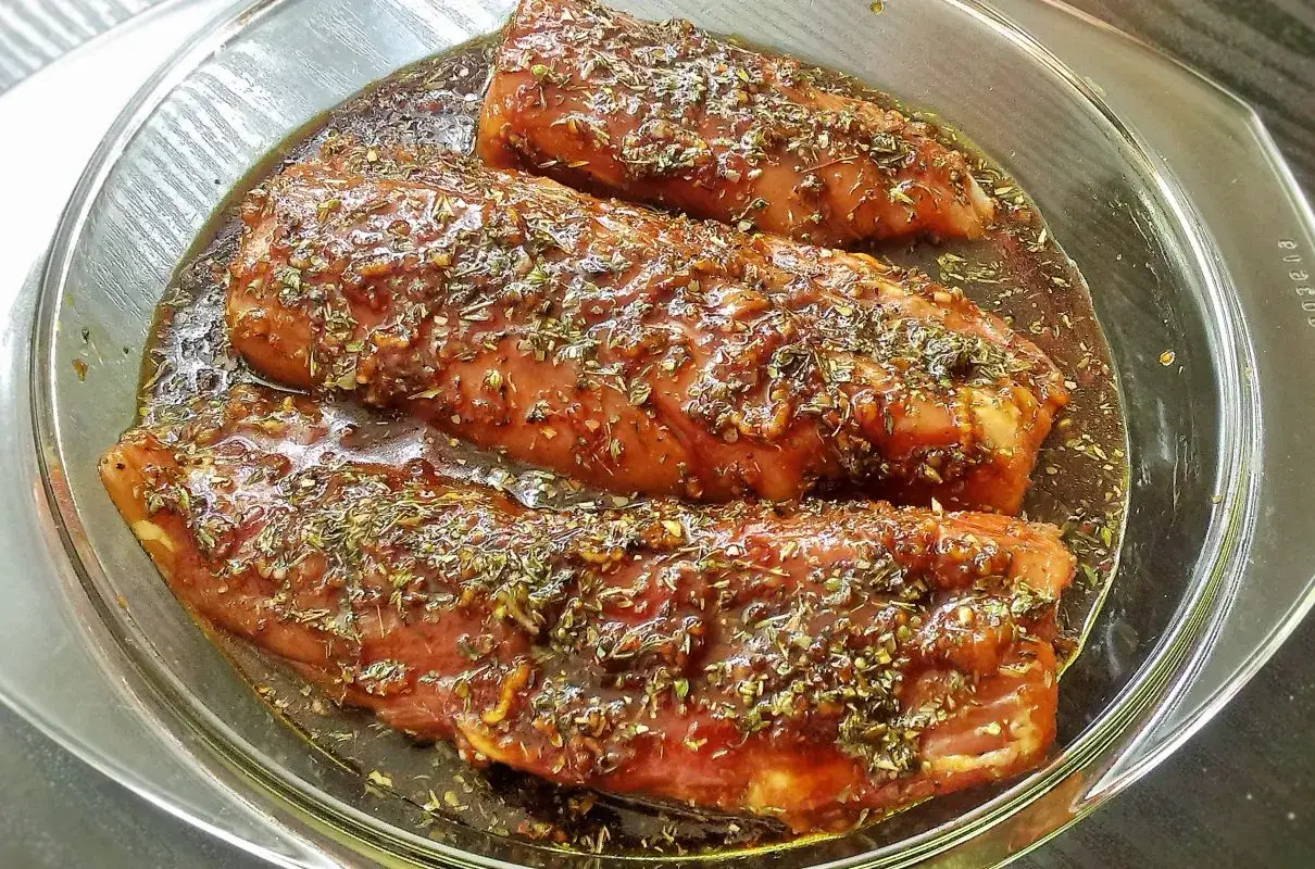 Trzy kawałki mięsa w szklanej misce, zanurzone w aromatycznej zalewie do mięsa z ziołami.