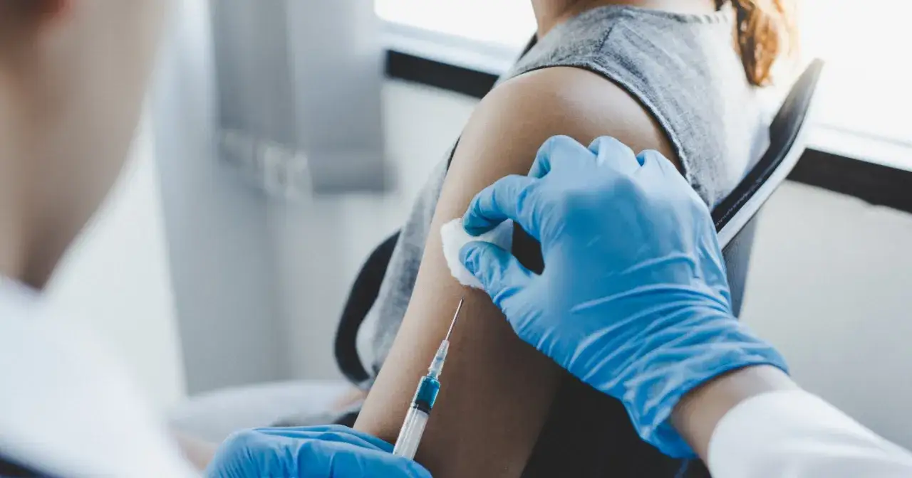 Ręka w niebieskiej rękawiczce podaje szczepienie na 18 lat. Wstrzyknięcie leku w ramię.