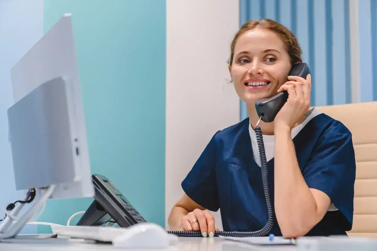 Uśmiechnięta recepcjonistka w granatowym uniformie odbiera telefon. Dzwoni, by dowiedzieć się, czy do dermatologa na NFZ trzeba mieć skierowanie.