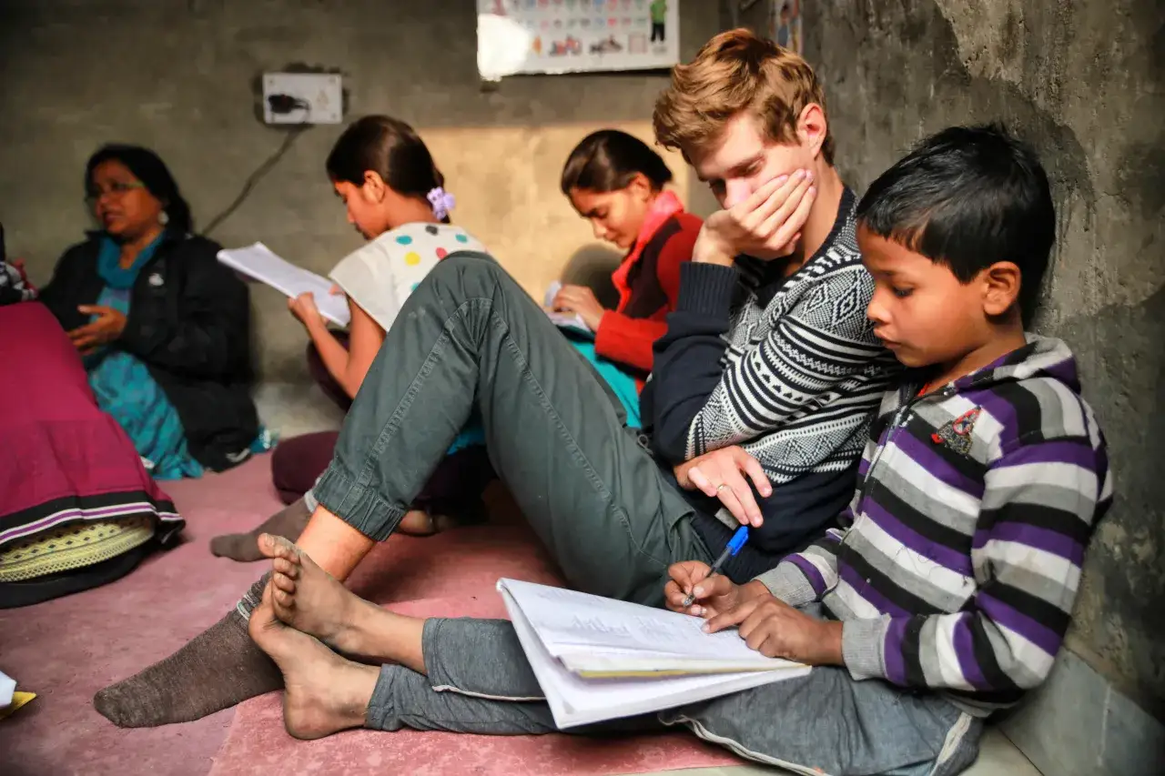 Dzieci uczą się w skromnej sali, która mogłaby być częścią największej szkoły na świecie. Młody wolontariusz pomaga im w nauce.