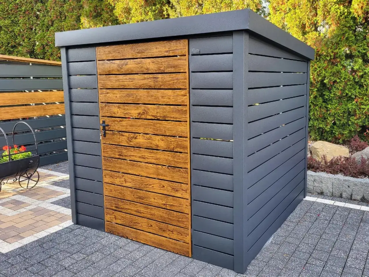 Nowoczesna wiata śmietnikowa przy domu, z drzwiami imitującymi drewno, dyskretnie ukrywa kosze.