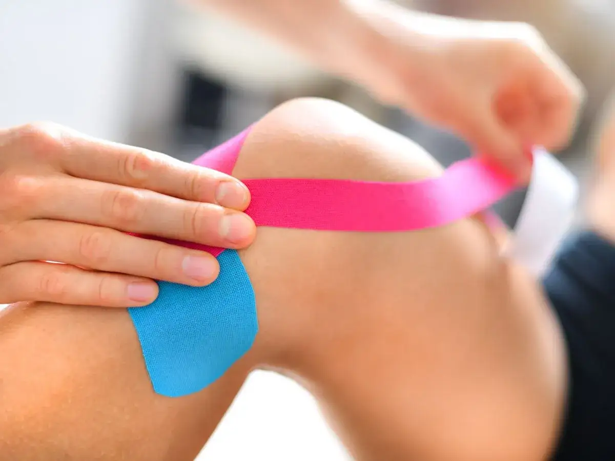 Dłonie przykładają różową i niebieską taśmę kinezjotapingową na kolano. Dowiedz się, jak przykleić tejpy na kolano, by zapewnić wsparcie.