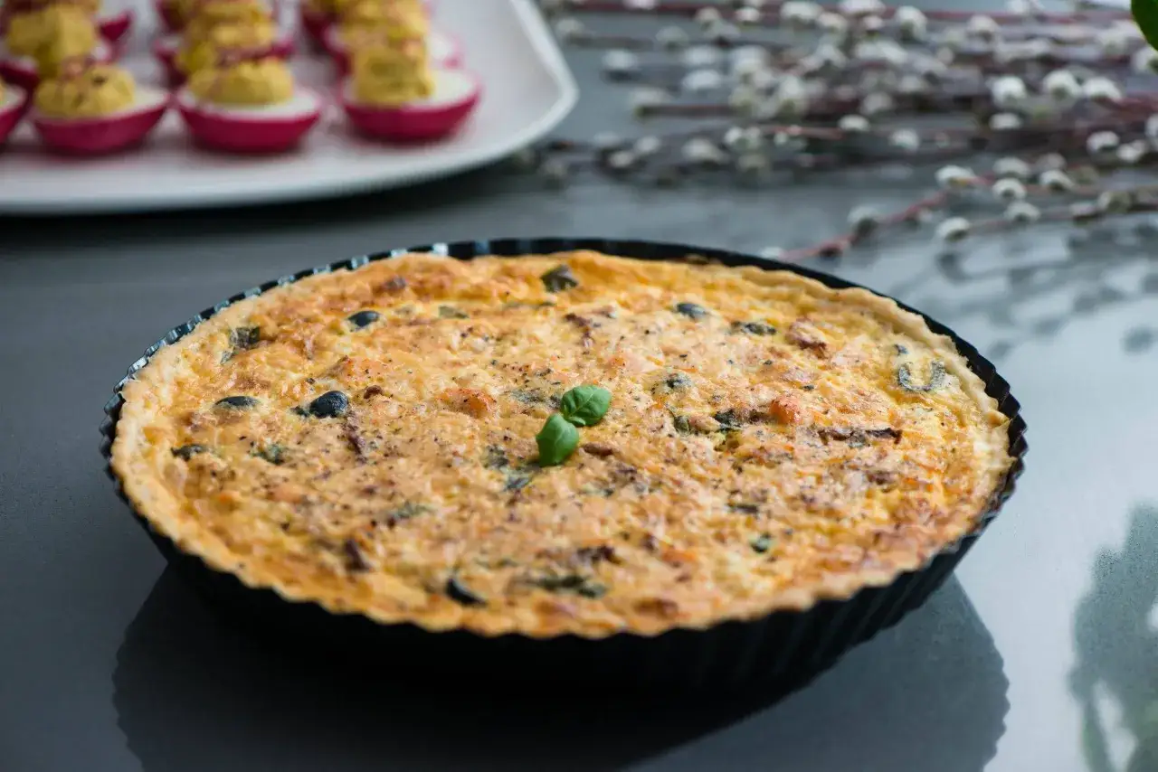 Złocista tarta z łososiem, udekorowana listkiem bazylii, kusi apetycznym wyglądem. Obok widać wielkanocne jajka faszerowane.