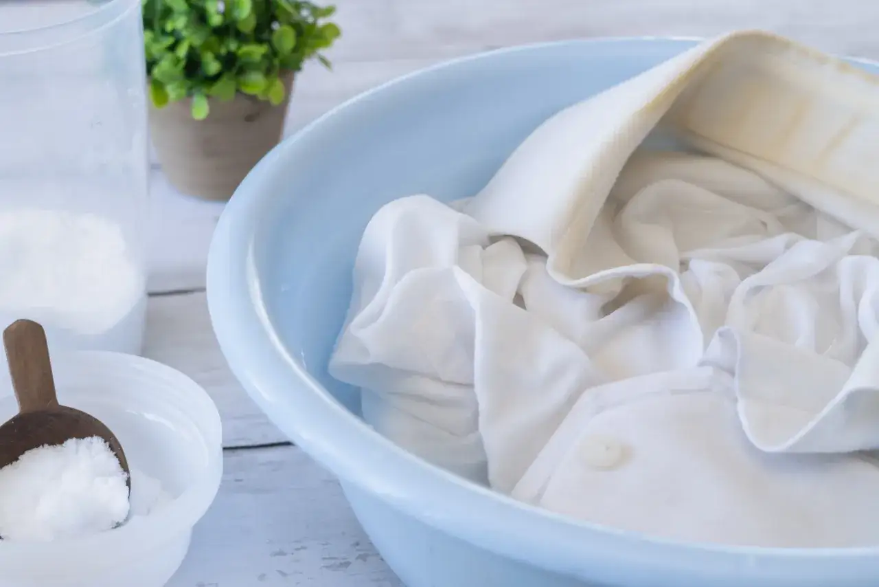 Biała bluzka z żółtymi plamami na kołnierzyku moczy się w niebieskiej misce. Obok proszek do prania, który pomoże jak sprać żółte plamy z białej bluzki.