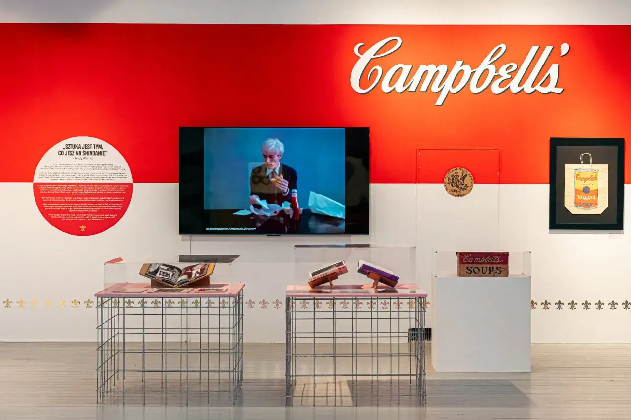 Wystawa prezentuje ruch artystyczny Andy'ego Warhola, z ekranem pokazującym artystę jedzącego kanapkę i logo Campbell's.
