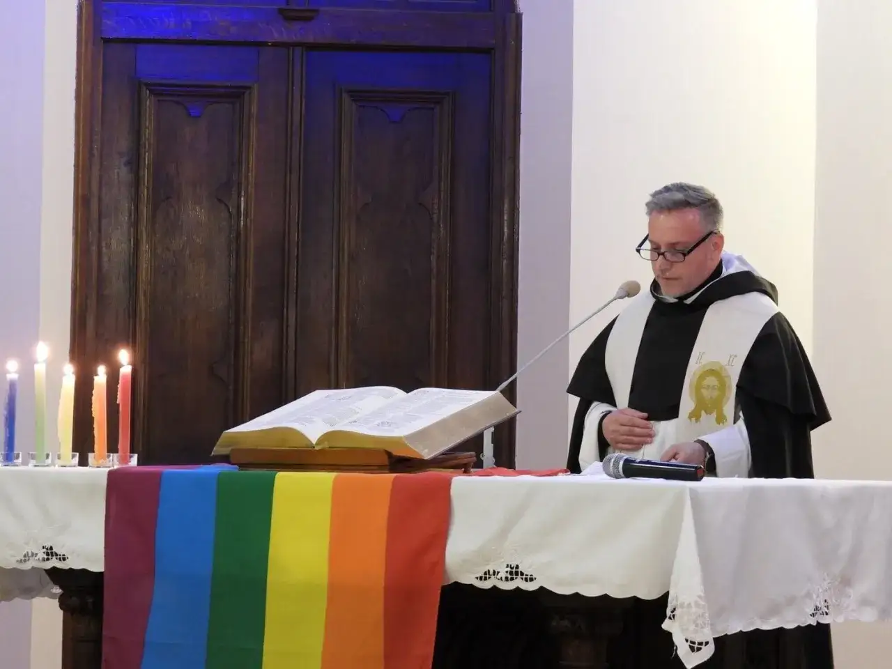 Duchowny w szacie z wizerunkiem Jezusa, przed tęczową flagą i otwartą Biblią. To pokazuje, co to znaczy pastor: otwartość i akceptacja.