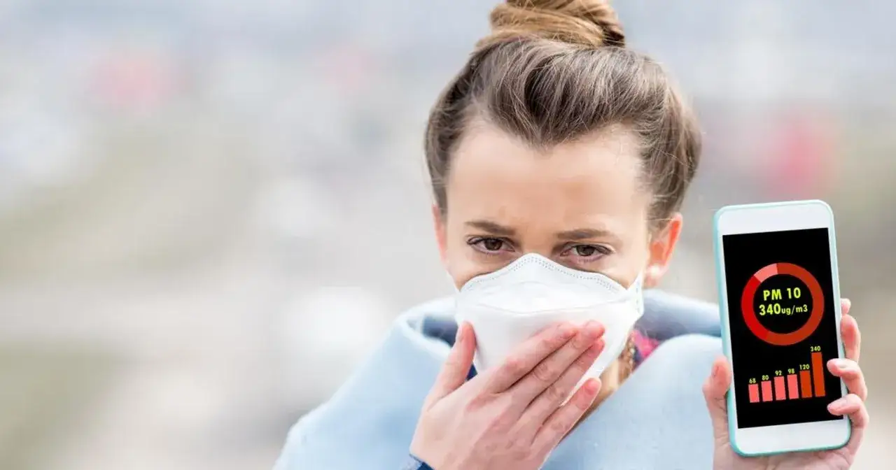 Kobieta w masce pokazuje telefon z aplikacją informującą o wysokim stężeniu pyłu PM10. Co to jest PM10? To szkodliwe cząstki w powietrzu.