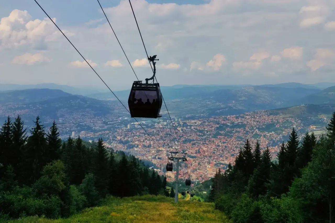 Kolejka linowa nad Sarajewem. Widok na miasto z góry, zalesione zbocze. Ceny biletów na kolejki mogą być różne.