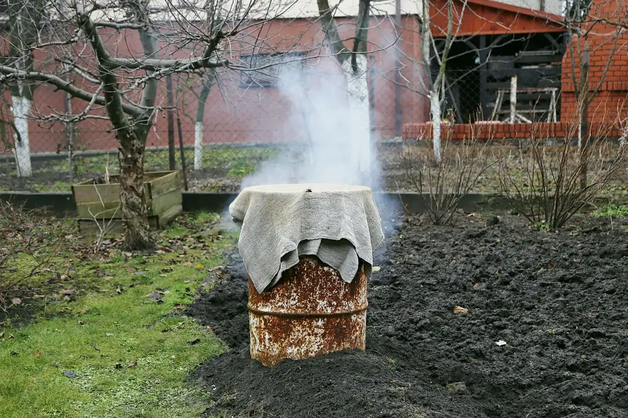 Dym unosi się z beczki, kt&oacute;ra służy jako prowizoryczna wędzarnia. Wok&oacute;ł niej ziemia i trawa.
