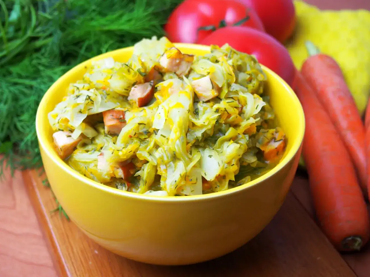 Pyszny bigos z młodej kapusty z kawałkami kiełbasy, podany w ż&oacute;łtej misce, obok marchewki i pomidor&oacute;w.