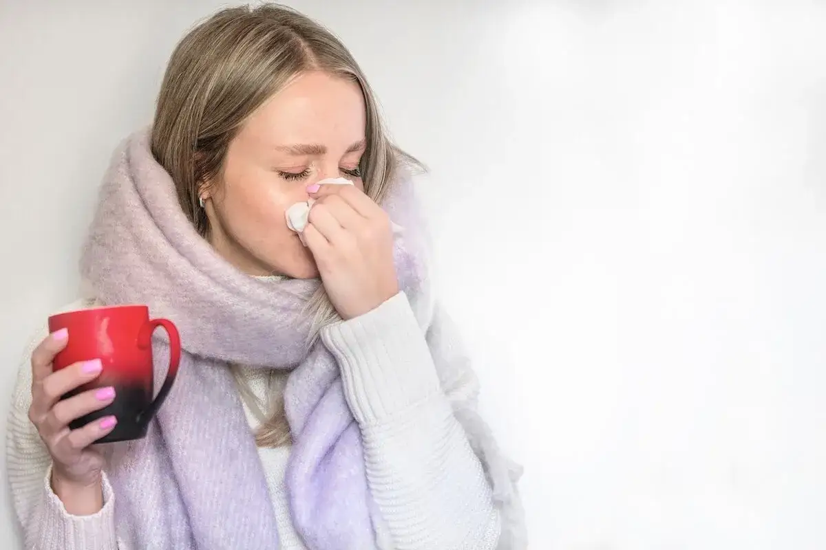 Kobieta z kubkiem w dłoni, wyciera nos chusteczką. Zmaga się z katarem, być może nawet z krwią.