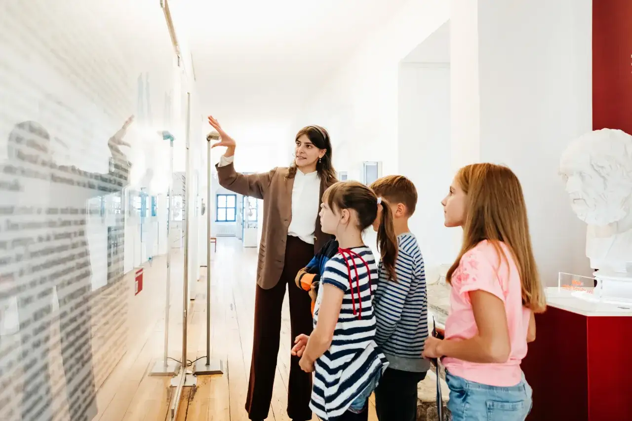 Nauczycielka oprowadza dzieci po muzeum. Czy rodzic może być opiekunem na wycieczce szkolnej? Dzieci słuchają z uwagą.