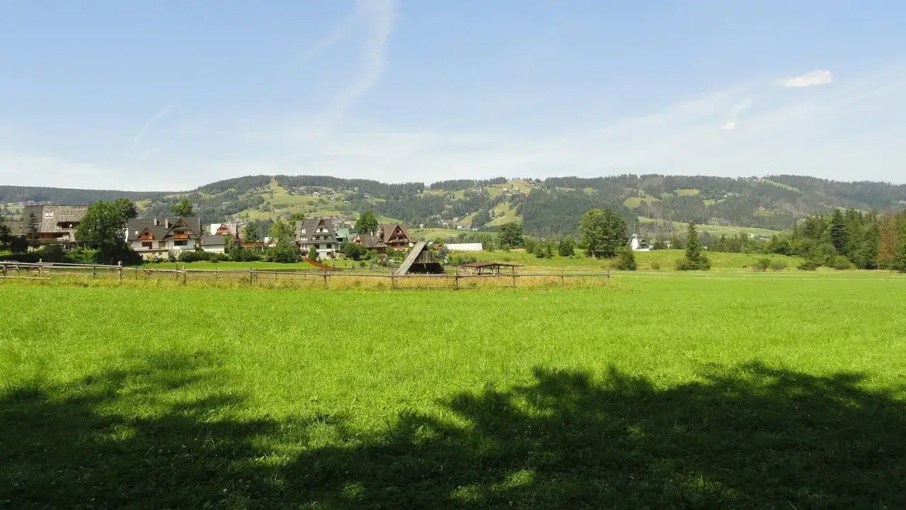 Zielona łąka z cieniem drzew, za nią drewniany płot i zabudowania. W tle góry i lasy, idealne na spacer ścieżką pod reglami.