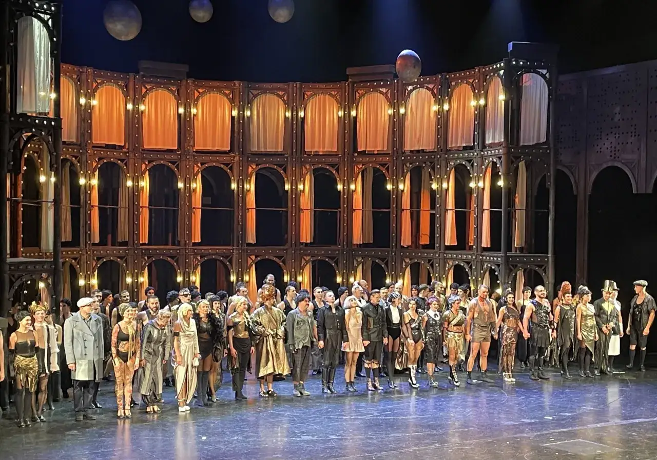Cała obsada teatru muzycznego na scenie, w barwnych strojach, na tle ozdobnej, wielopoziomowej scenografii z kurtynami i światłami.
