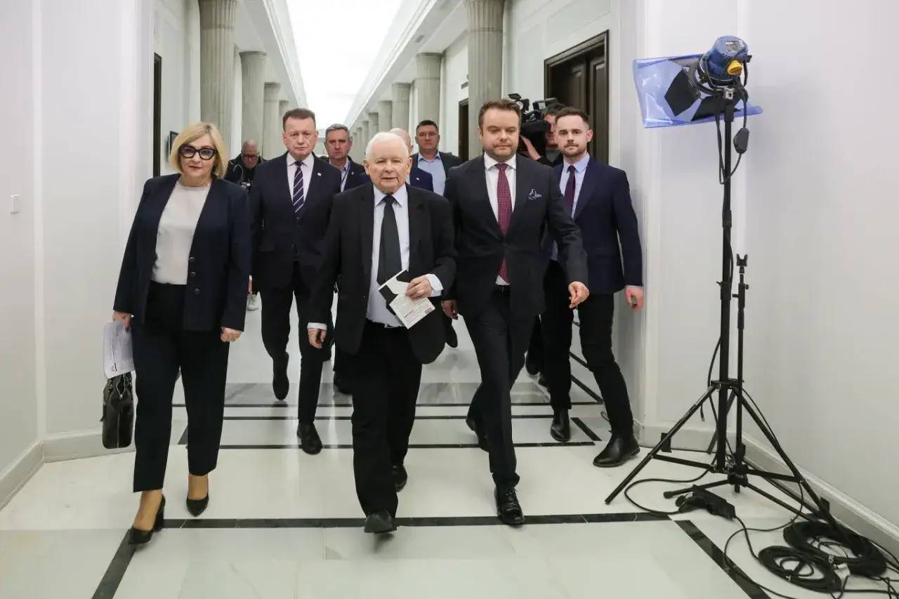 Jarosław Kaczyński i inni politycy idą korytarzem. W tle widać kamery i sprzęt oświetleniowy, co może sugerować okres, w jakich latach rządził PiS.