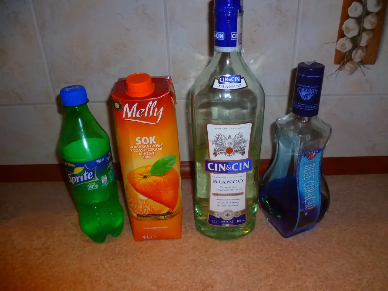 Na stole stoi butelka Sprite, karton soku pomarańczowego Melly i dwie butelki alkoholu: Cin&Cin Bianco i Blue Curacao. Idealny zestaw na drinki z Żabki!