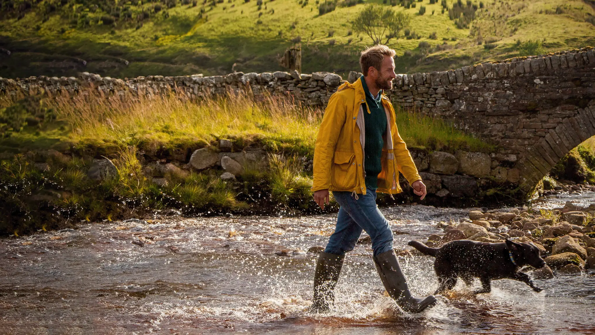 Mężczyzna w żółtej kurtce i pies przemierzają strumień. W tle kamienny most i zielone wzgórza. Witamy na odludziu.