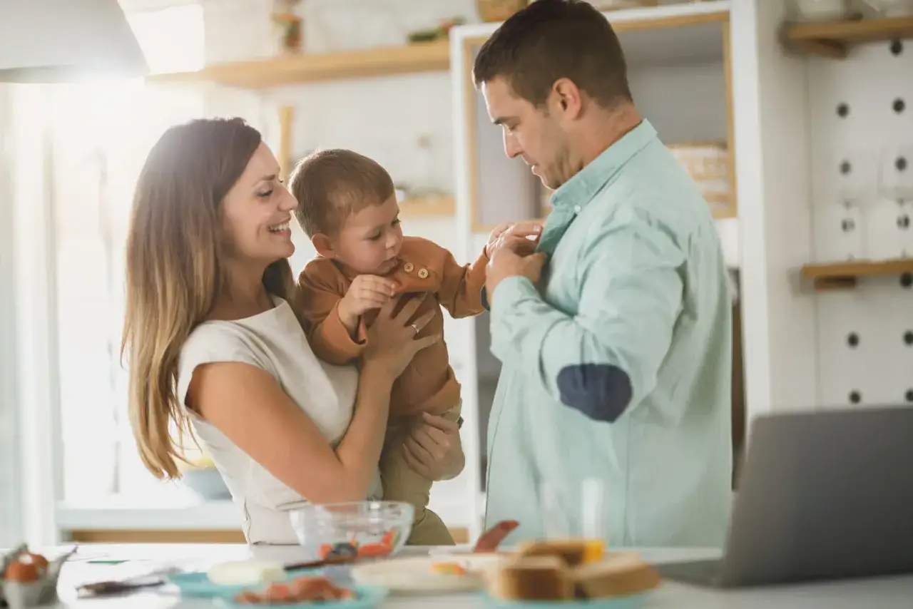Rodzina w kuchni. Dziecko w ramionach mamy, ojciec zapina mu koszulę. Pamiętaj, czego nie można robić po szczepieniu niemowlaka, by zapewnić mu komfort.