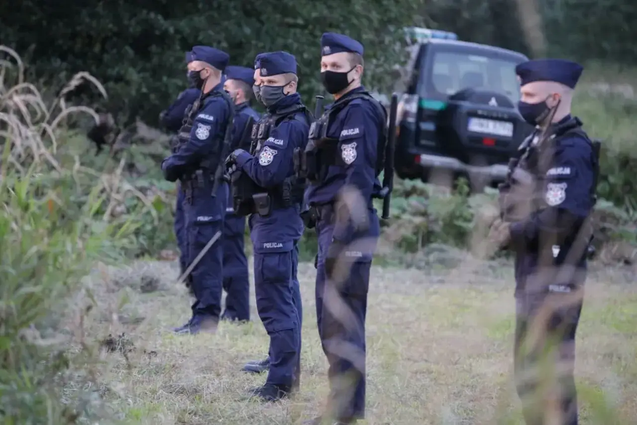 Policjanci w mundurach i maseczkach stoją w gotowości. W tle widać radiowóz. Sytuacja wygląda na stan wyjątkowy.
