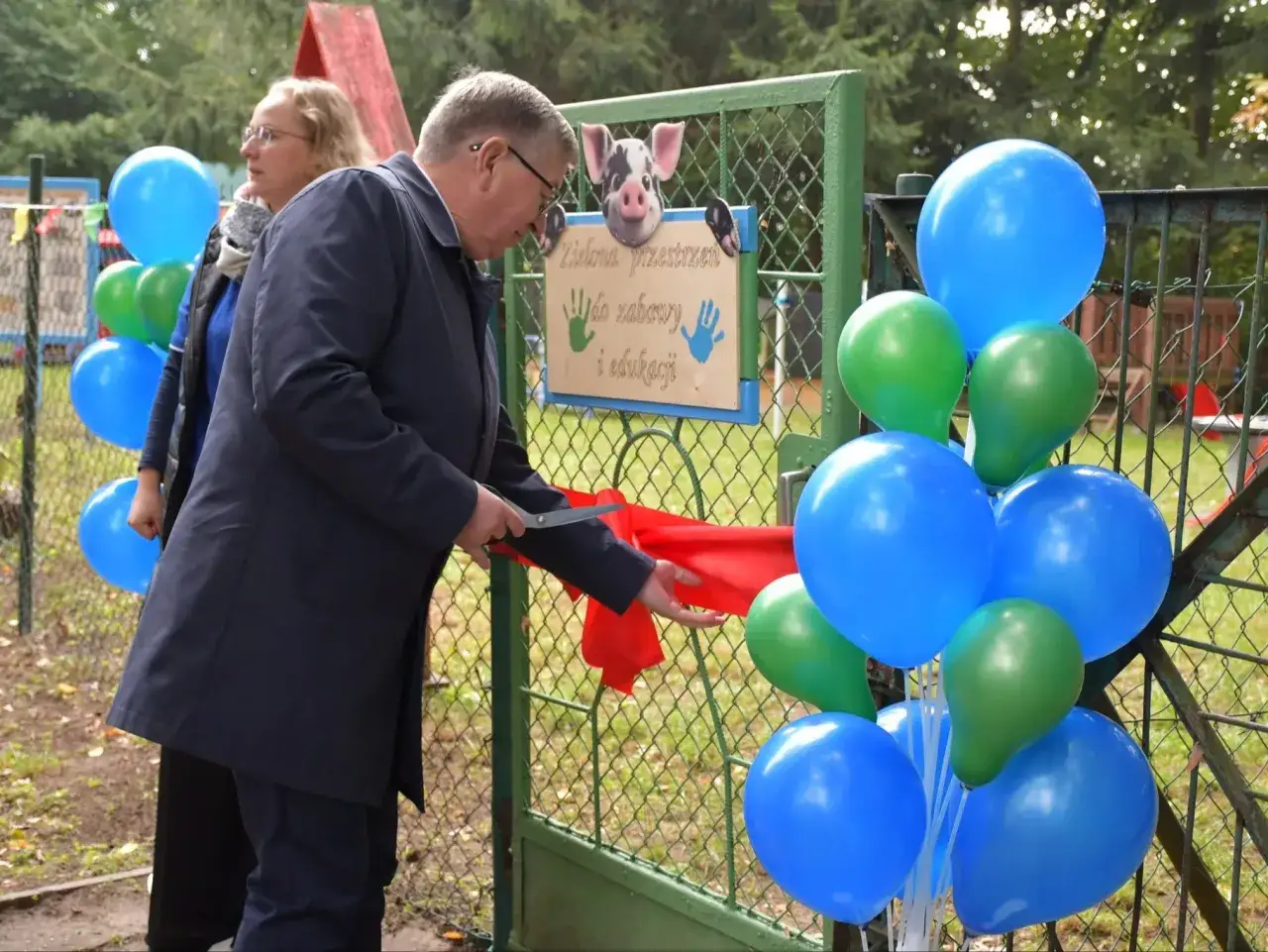 Uroczyste otwarcie placu zabaw dla dzieci. Pan przecina wstęgę, balony w kolorach niebieskim i zielonym.