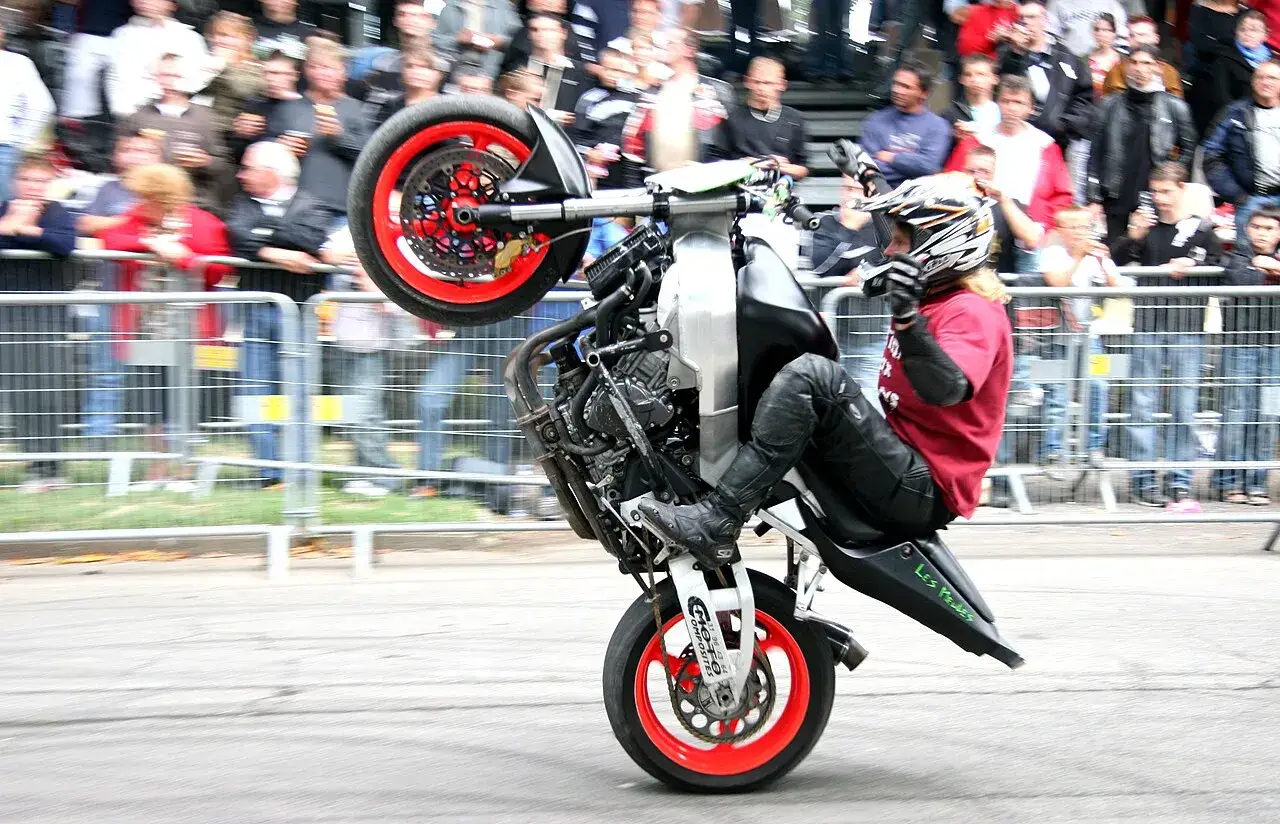 Motocykl do stuntu na tylnym kole, kierowca w kasku i kombinezonie, tłum widz&oacute;w za barierkami.