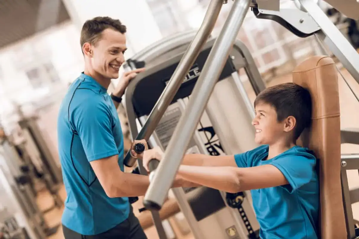 Ojciec i syn ćwiczą razem na siłowni. Chłopiec, który ma 13 lat, z uśmiechem korzysta z maszyny do ćwiczeń.