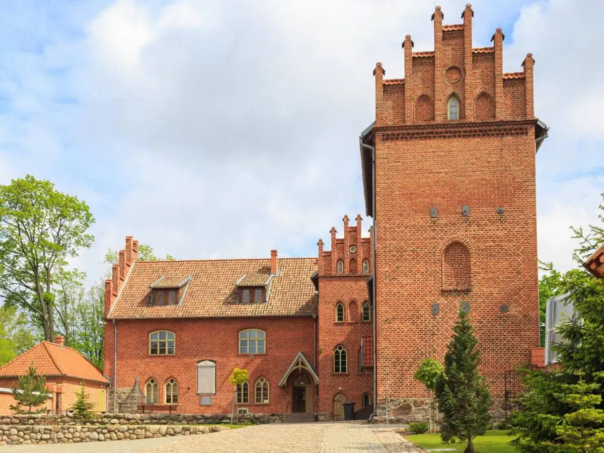 Zamek w Grunwaldzie, ceglana wieża i budynki z czerwonej cegły, otoczone zielenią.