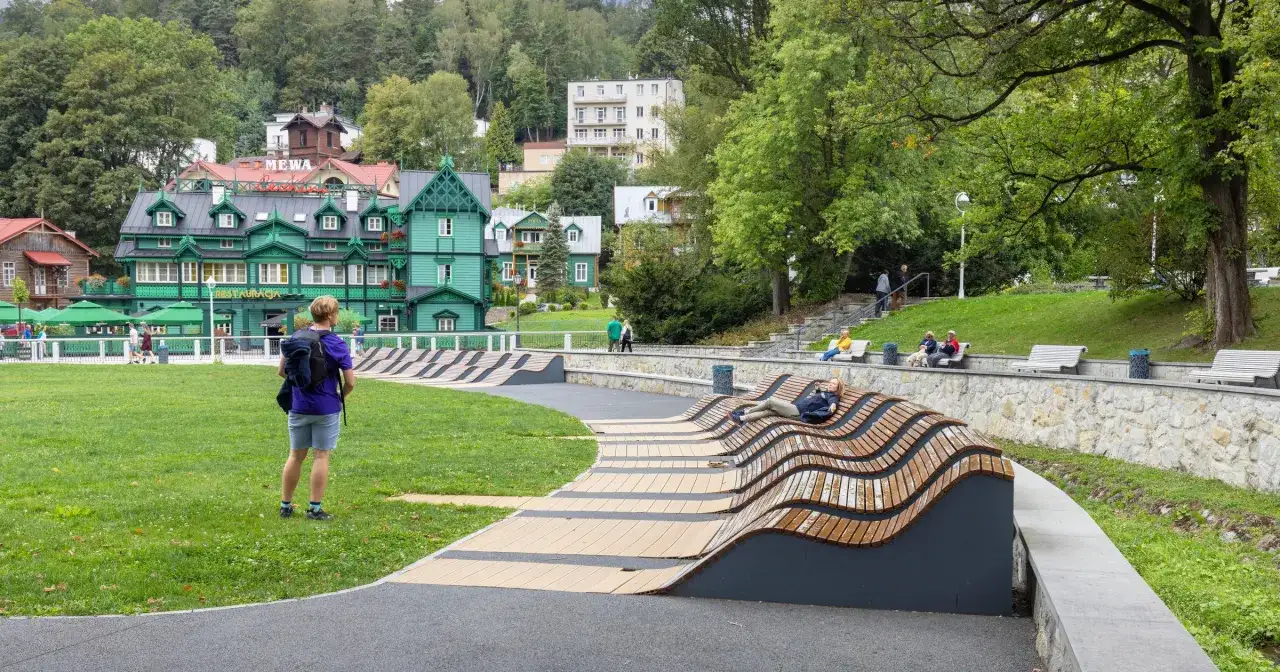 Zielony budynek sanatorium w Krynicy-Zdroju, z widokiem na park i turyst&oacute;w. Sprawdź sanatorium Krynica Zdr&oacute;j cennik.