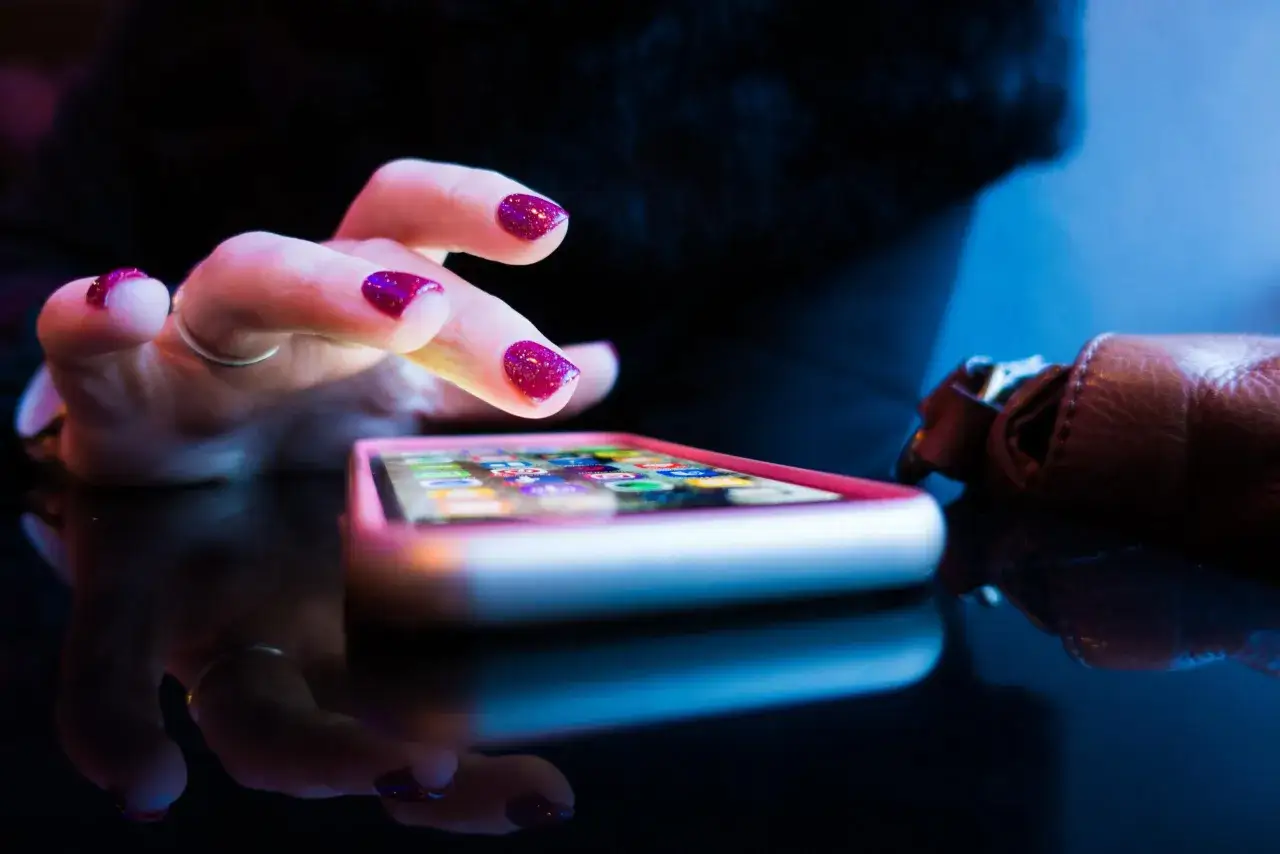 Dłoń z błyszczącymi paznokciami dotyka ekranu telefonu, na kt&oacute;rym widać ikony aplikacji do nauki polskiego.