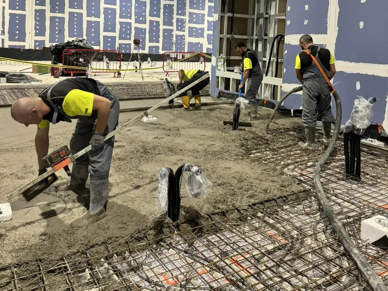 Pracownicy wylewają beton na zbrojenie. Pokazują, jak zrobić wylewkę betonową, równając ją łopatami i niwelatorami.