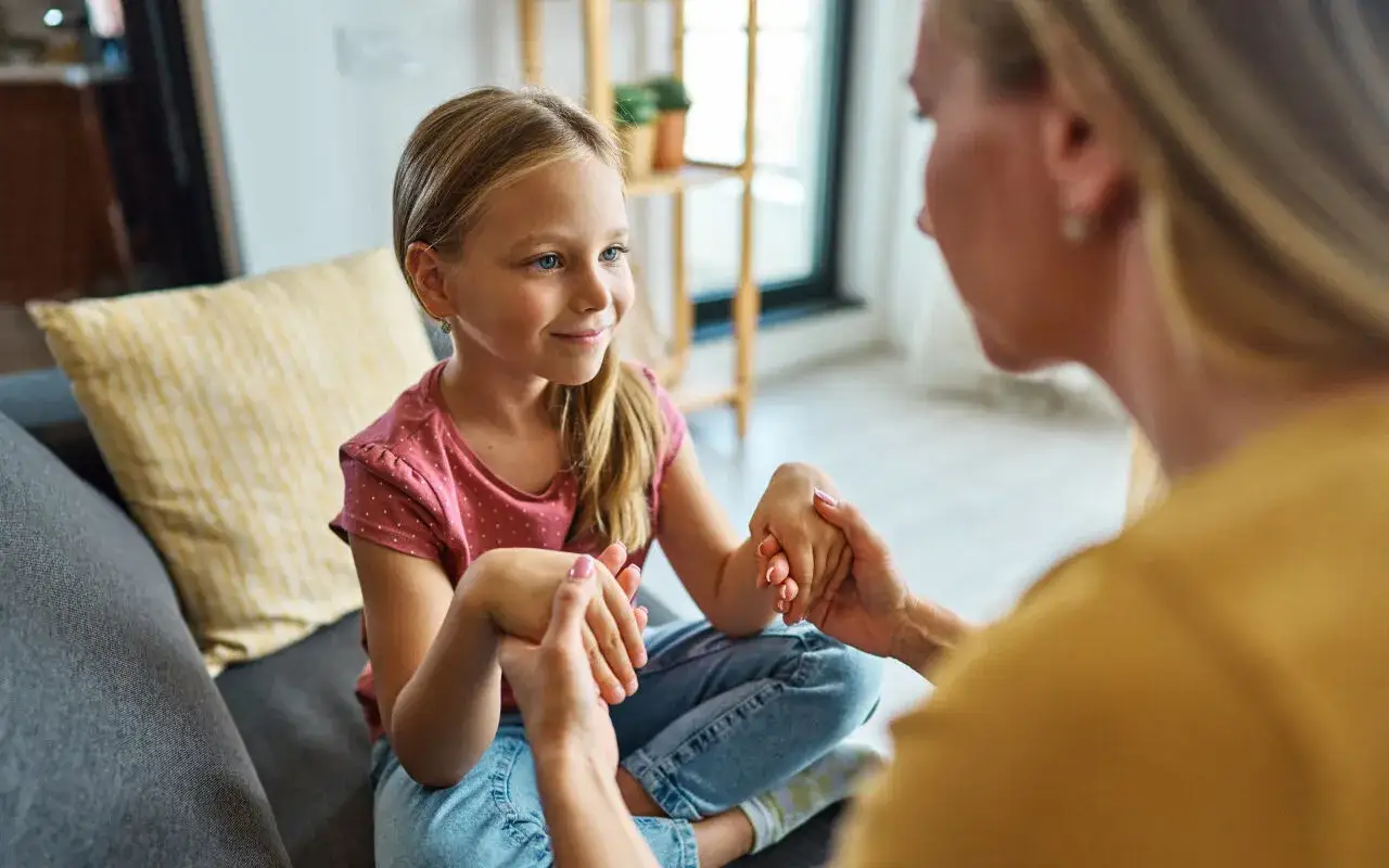 Mama spokojnie tłumaczy córce, jak rozmawiać z dzieckiem o złym zachowaniu. Dziewczynka słucha uważnie, trzymając dłonie mamy.