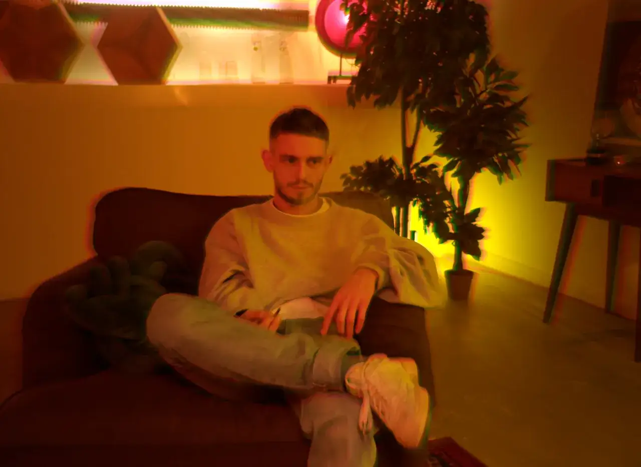 DJ BRK robię co mogę, siedząc na kanapie w przytulnym pokoju z rośliną i geometrycznymi ozdobami na ścianie.