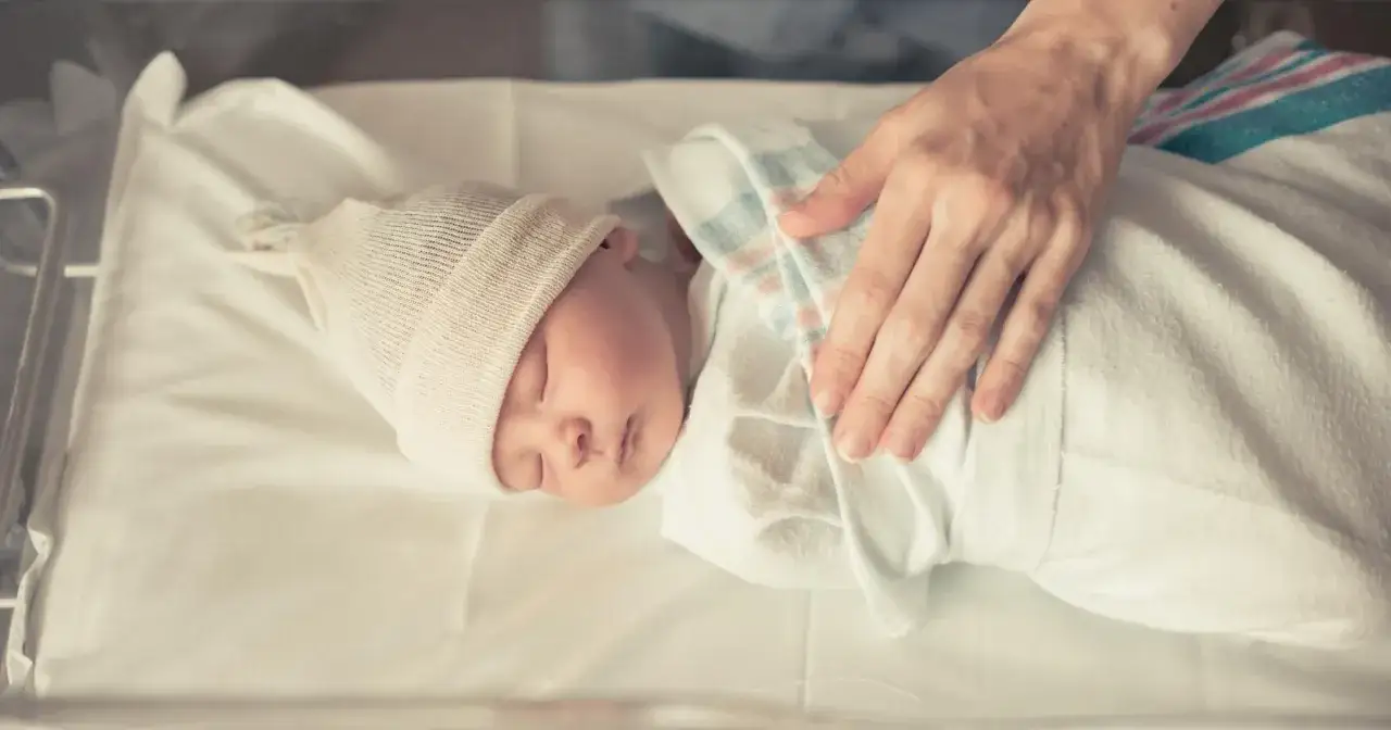 Dziecko urodzone w 37 tygodniu nie jest uznawane za wcześniaka. Maluch śpi w ł&oacute;żeczku, otulony kocykiem, a ręka opiekuna delikatnie go głaszcze.