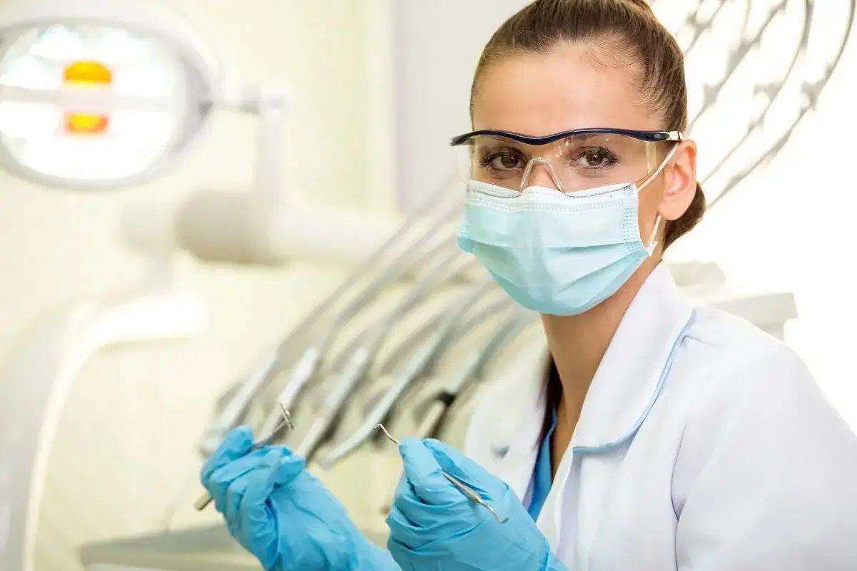 Dentystka w maseczce i okularach ochronnych, z rękawiczkami na dłoniach, trzyma narzędzia. Odpowiedź na pytanie: czy u dentysty trzeba mieć maseczkę? Tak, dla bezpieczeństwa.