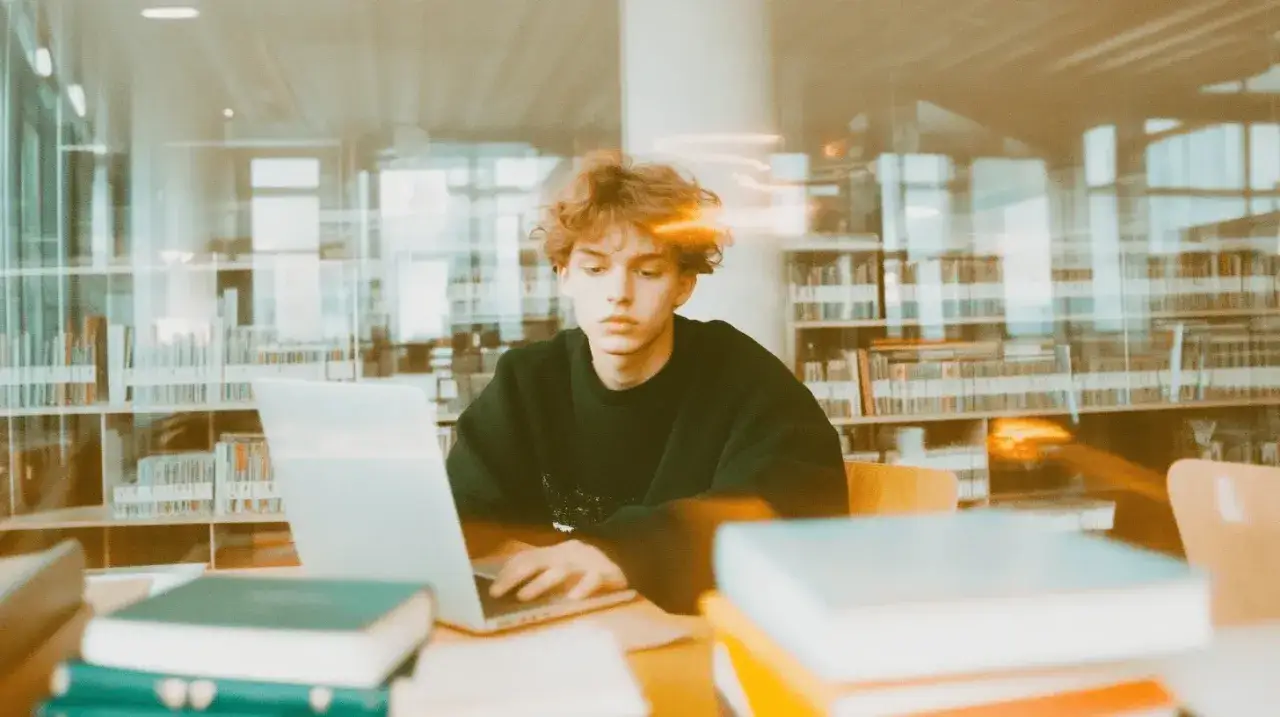 Młody mężczyzna z kręconymi włosami pracuje na laptopie w bibliotece. W tle widać regały pełne książek. credits po polsku.