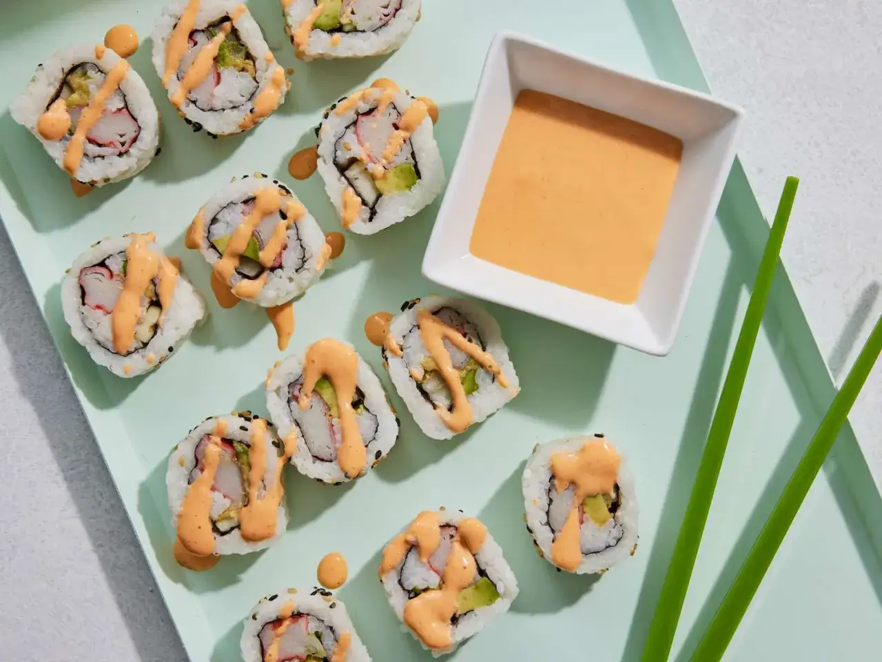 Roladki sushi z awokado i krabem, polane ostrym dodatkiem do sushi. Obok miseczka z sosem i pałeczki.