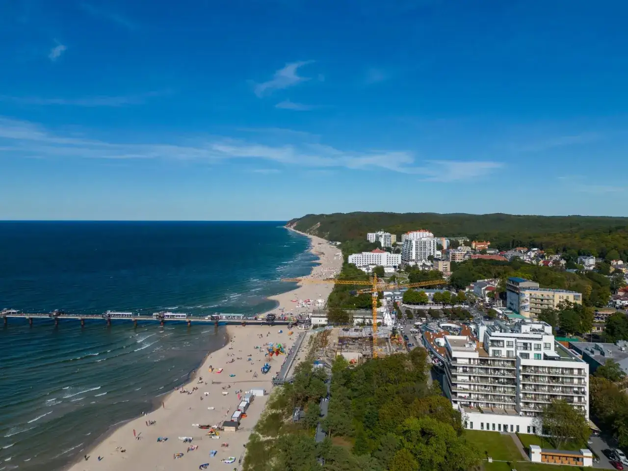 Widok na plażę, molo i budynki nad morzem. W oddali widać las, a bliżej nowoczesne obiekty, w tym potencjalne sanatorium dla dzieci nad morzem.