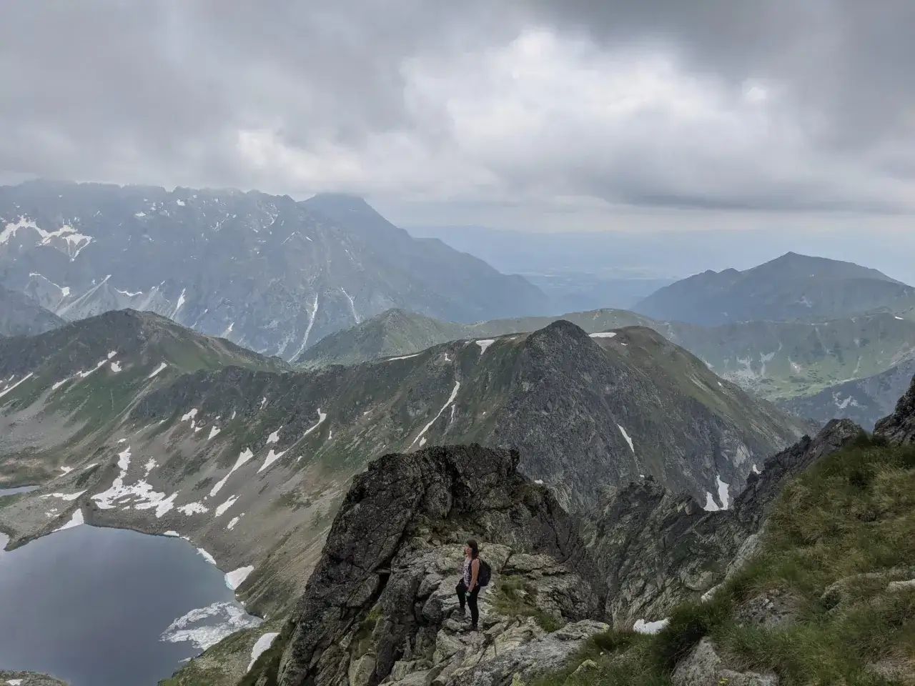 Widok na Tatry z turystką na skale. Czy szlak na Świnicę jest otwarty? W oddali jezioro i kolejne pasma g&oacute;rskie pod zachmurzonym niebem.