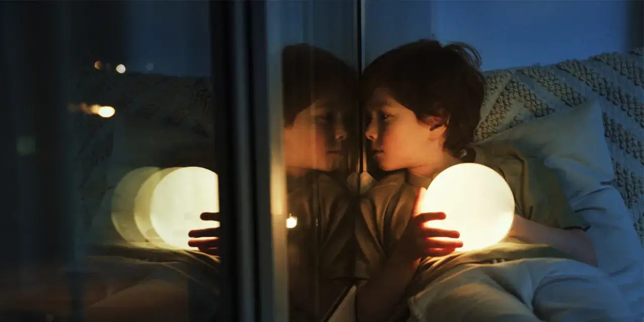 Chłopiec trzymający świecącą kulę, zastanawiający się, dlaczego dziecko lunatykuje. Jego odbicie w szybie wygląda jak sen.