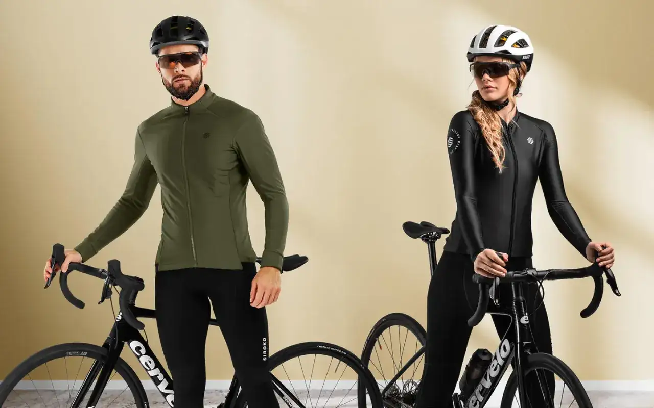 Para idealnego **bike fitting**: mężczyzna w zielonej bluzie i kobieta w czarnej, oboje w kaskach i okularach, pozują z rowerami.