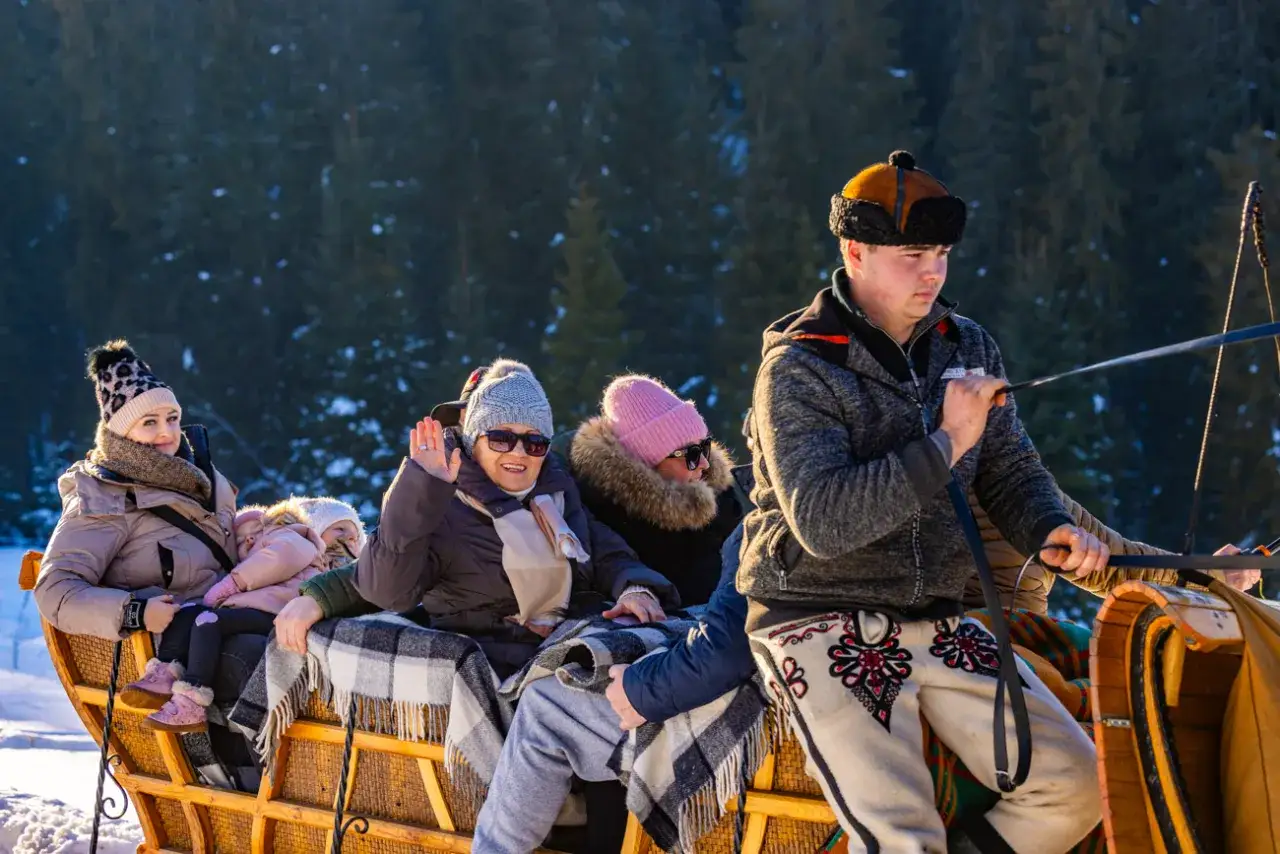 Rodzina cieszy się zimowym kuligiem w Bukowinie Tatrzańskiej. Dzieci i dorośli otuleni kocami, uśmiechnięci, podr&oacute;żują saniami po ośnieżonym lesie.
