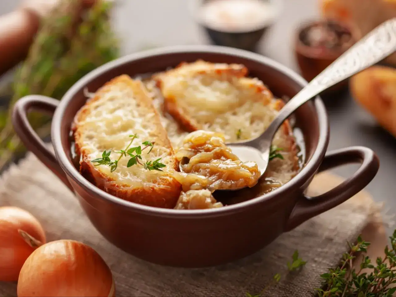 Aromatyczna zupa cebulowa z grzankami zapieczonymi serem, podana w brązowej miseczce. Na łyżce widoczna porcja zupy z cebulą i grzanką.