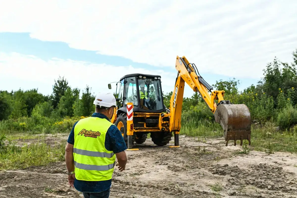 Operator w kamizelce odblaskowej i kasku patrzy na koparkę. Dowiedz się, ile kosztują uprawnienia na koparki, aby rozpocząć karierę.