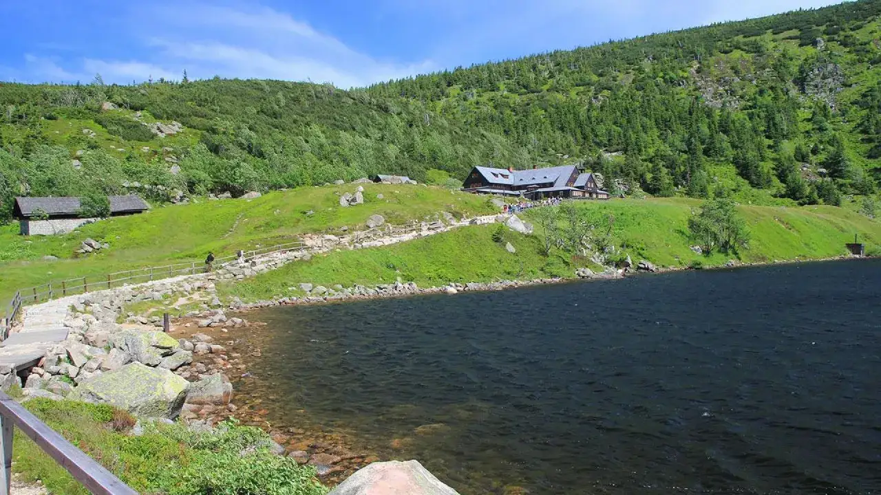 Kamienista ścieżka prowadzi do schroniska na tle zielonych zboczy, gdzie widać najwyższe szczyty Karkonoszy.