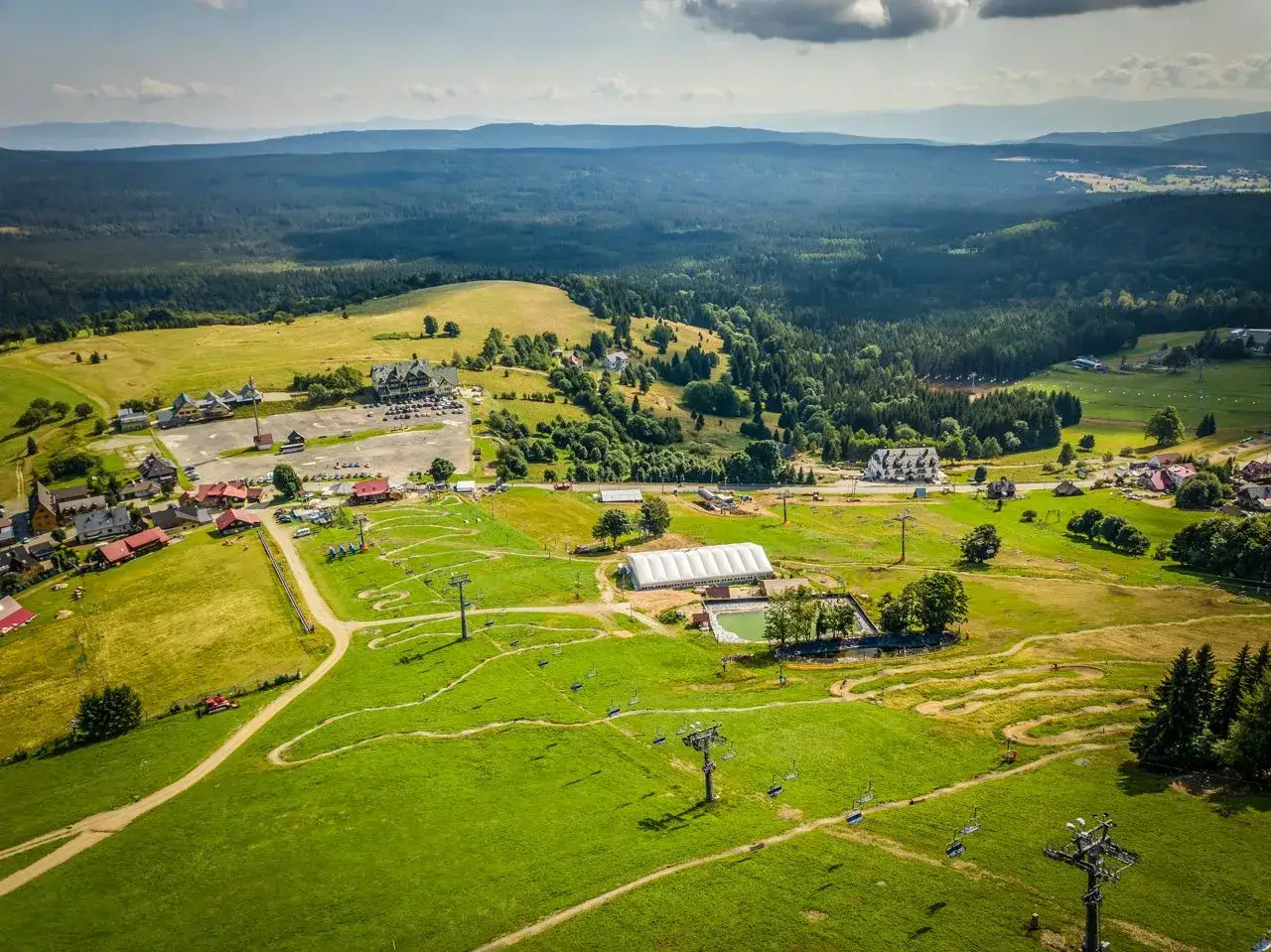 Zieleniec bike park w letniej odsłonie. Widok z lotu ptaka na zielone wzgórza, wyciągi krzesełkowe, tor rowerowy i budynki ośrodka.