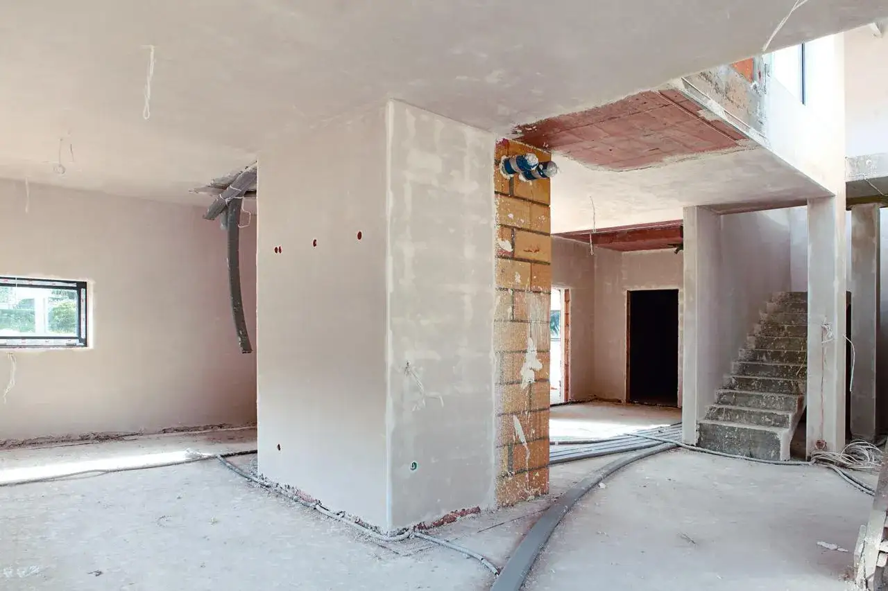 Budowa domu w toku. Widać ściany z tynkiem, instalacje elektryczne i schody. Trudno ocenić, ile cm tynku na ścianie, ale prace postępują.